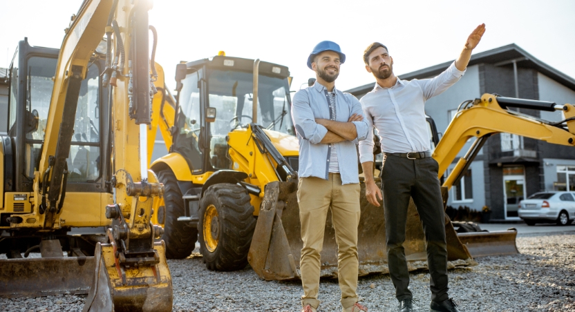 Two men wearing hard hats stand beside construction equipment, pointing into the distance, symbolizing strategic planning and partnership in the equipment sales industry.